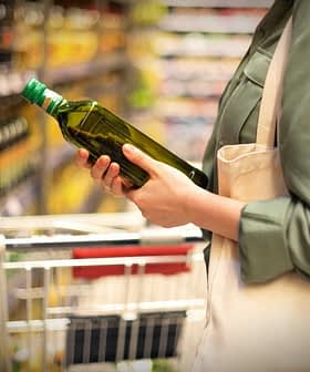 Individual holding a green bottle of olive oil while shopping in a grocery store aisle. - Olive Oil Times