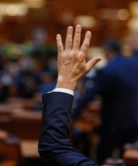 Uma mão levantada em um ambiente formal, possivelmente durante uma votação em uma assembleia legislativa. - Olive Oil Times