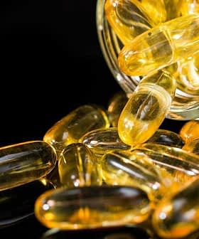 Un frasco de vidrio volcado con cápsulas de gel amarillas derramándose sobre una superficie reflectante. Olive Oil Times