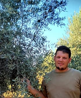 Moški, ki stoji ob oljkah na prostem, z zelenim listjem v ozadju. - Olive Oil Times