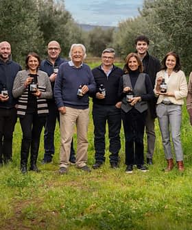 Gruppo di dodici individui in piedi in un uliveto, ognuno con in mano una bottiglia di olio d'oliva. Olive Oil Times