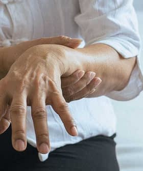 Individual with a white shirt holding their wrist while seated on a bed. - Olive Oil Times