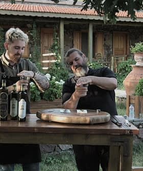 Dvojica muškaraca koji sudjeluju u kušanju maslinovog ulja za drvenim stolom na otvorenom. - Olive Oil Times