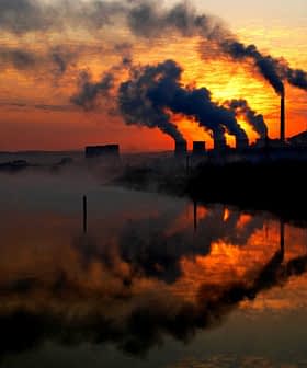 Chaminés industriais silhuetadas emitindo fumaça contra um céu de pôr do sol com reflexos na água. - Olive Oil Times