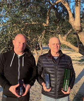 Zeytin ağacının önünde, ellerinde zeytinyağı şişeleri tutan iki adam dışarıda duruyor. - Olive Oil Times
