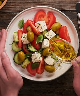 Mãos derramando azeite de oliva sobre uma salada com tomates, pepinos, azeitonas e queijo feta em um prato. - Olive Oil Times
