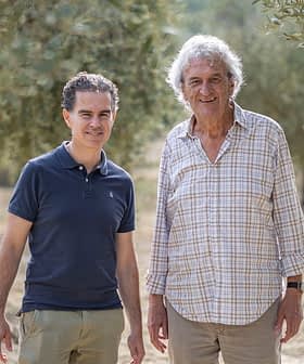 Δύο άντρες στέκονται σε έναν ελαιώνα, χαμογελαστοί και ντυμένοι πρόχειρα με ελαφριά ρούχα. - Olive Oil Times