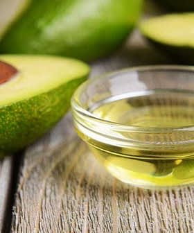 Narezan avokado s koščico poleg majhne steklene posode z avokadovim oljem na leseni površini. - Olive Oil Times