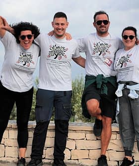 Açık havada, aynı tişörtleri giymiş ve kameraya gülümseyen dört kişi birlikte poz veriyor. - Olive Oil Times