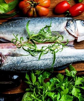 Dos pescados crudos colocados en una tabla de cortar de madera rodeados de verduras y hierbas frescas. Olive Oil Times