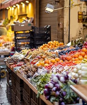 - Pazar ortamında arabalarla ve kasalarla çeşitli meyve ve sebzelerin canlı bir şekilde sergilenmesi. Olive Oil Times