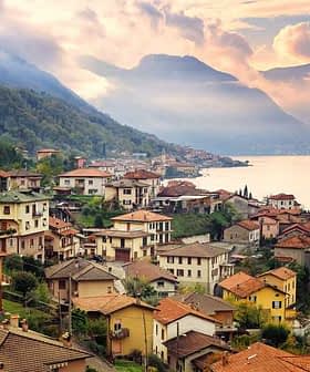 Villaggio sul lago con case e montagne sullo sfondo sotto un cielo nuvoloso. - Olive Oil Times