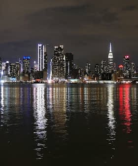 Vista noturna do horizonte da cidade de Nova York com edifícios iluminados refletindo na água. - Olive Oil Times