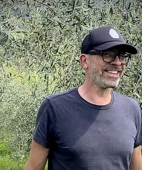 Un homme portant une casquette noire et des lunettes souriant debout dans une oliveraie. - Olive Oil Times