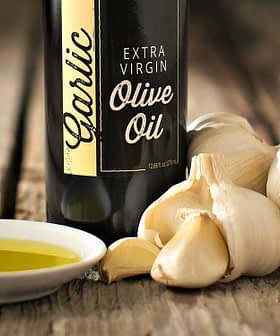 Bottiglia di olio extravergine di oliva all'aglio accanto a bulbi d'aglio e un piattino di olio d'oliva. Olive Oil Times