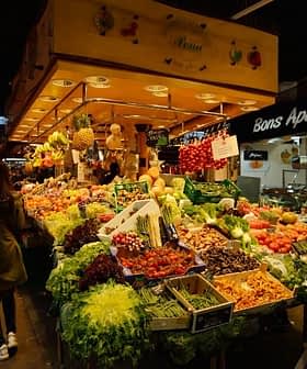 Ένας πολυσύχναστος πάγκος αγοράς με ποικιλία φρούτων και λαχανικών, όπως χόρτα, ντομάτες και σταφύλια. - Olive Oil Times