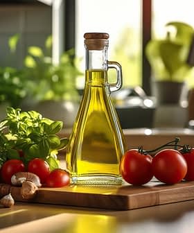 Eine Glasflasche Olivenöl, umgeben von frischen Tomaten und Basilikum auf einem Holzschneidebrett. – Olive Oil Times