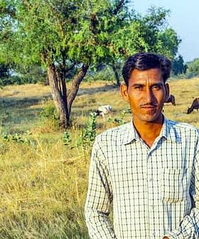 Un uomo con una camicia a quadri è in piedi in un campo con del bestiame al pascolo sullo sfondo. - Olive Oil Times