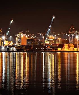 Visão noturna de um porto com contêineres iluminados e guindastes refletindo na água. - Olive Oil Times