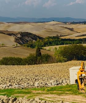 Gele tractor geparkeerd bij een schuur in een landelijk agrarisch landschap met glooiende heuvels. Olive Oil Times