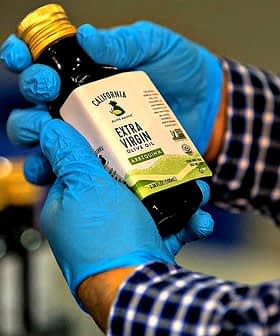 Osoba u plavim rukavicama drži bocu kalifornijskog ekstra djevičanskog maslinovog ulja s etiketom. - Olive Oil Times