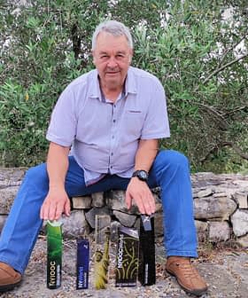 Un hombre sentado en un muro de piedra con varias botellas de aceite de oliva expuestas frente a él. Olive Oil Times