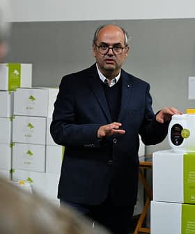 Een man in pak presenteert een fles olijfolie in een pakhuis. Olive Oil Times