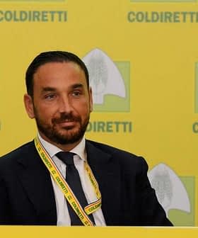 Man in pak en stropdas zit aan een tafel met een Coldiretti-banner op de achtergrond. Olive Oil Times