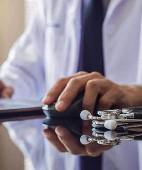 Um profissional médico de jaleco branco usando um laptop com um estetoscópio sobre a mesa. - Olive Oil Times