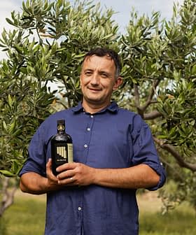 Čovjek stoji u masliniku i drži bocu maslinova ulja s maslinama u pozadini. - Olive Oil Times