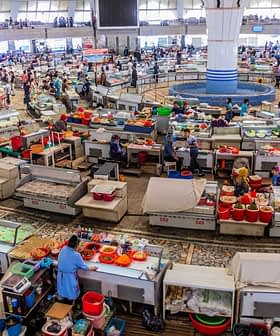 Um mercado movimentado, cheio de vendedores e clientes, exibindo diversas barracas e produtos em um grande espaço interno. - Olive Oil Times