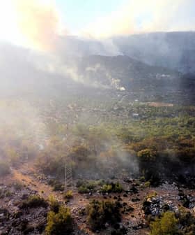 Vue aérienne d'un paysage avec de la fumée s'élevant du sol parmi les arbres et le terrain rocheux. - Olive Oil Times