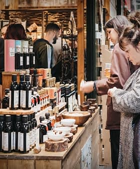 Verschillende flessen olijfolie staan ​​uitgestald op een houten tafel op een markt, met mensen op de achtergrond. Olive Oil Times