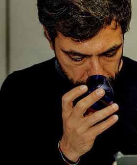 黒髪とあごひげの男性が青いガラスのコップを顔の近くに持っています。 - Olive Oil Times