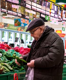 Un homme âgé examine des légumes dans une épicerie avec des bacs à produits verts. Olive Oil Times
