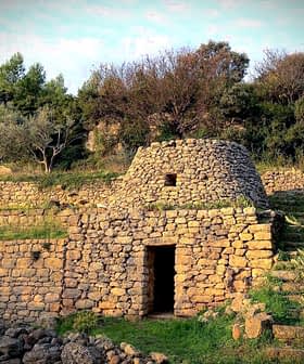 Μια πέτρινη κατασκευή με κωνική στέγη που περιβάλλεται από πράσινο και πέτρινους τοίχους. - Olive Oil Times