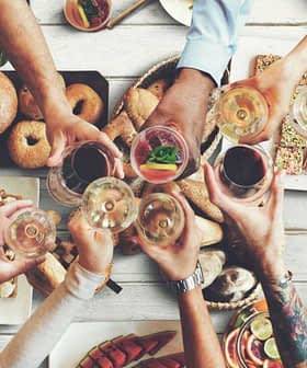 Grupo de personas brindando con copas sobre una mesa llena de diversos alimentos y bebidas. - Olive Oil Times