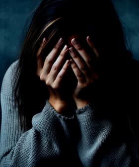 Individuo con cabello largo y oscuro cubriéndose la cara con las manos, vistiendo un suéter gris sobre un fondo oscuro. - Olive Oil Times