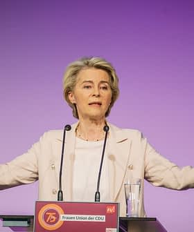 Ženska gestikulira med govorom na odru med dogodkom z vijoličnim ozadjem. - Olive Oil Times