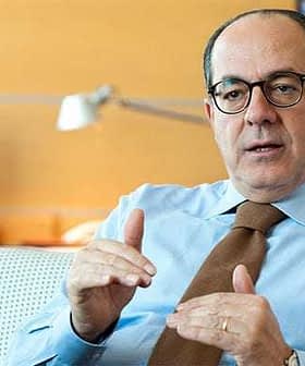 Επιχειρηματίας που φορά ένα γαλάζιο πουκάμισο και γυαλιά, χειρονομώντας ενώ κάθεται σε ένα περιβάλλον γραφείου. - Olive Oil Times