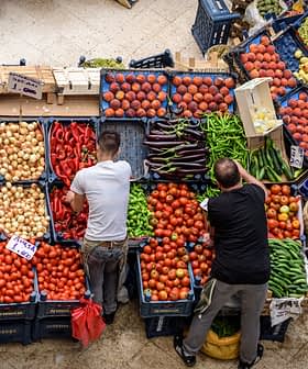 Zwei Personen arrangieren frisches Gemüse und Obst an einem Marktstand mit verschiedenen Produkten. - Olive Oil Times