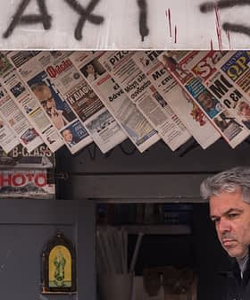 Un hombre de pie frente a un quiosco que exhibe varios periódicos y revistas. - Olive Oil Times
