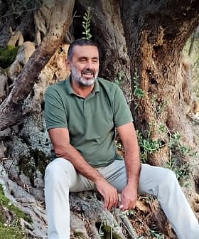 Mann mit Bart, der auf den Wurzeln eines Olivenbaums sitzt und ein grünes Hemd und eine helle Hose trägt. - Olive Oil Times