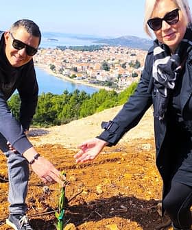 Δύο άτομα σκύβουν για να φυτέψουν ένα μικρό δέντρο σε μια αγροτική περιοχή με θέα σε μακρινούς λόφους και το νερό. - Olive Oil Times