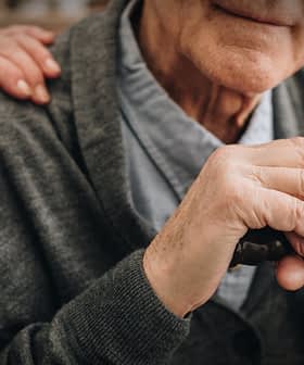 Close das mãos de um homem idoso apoiadas em uma bengala, com uma mão em seu ombro. - Olive Oil Times