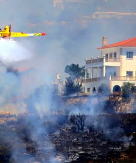 Avión de bomberos amarillo liberando agua sobre un incendio forestal cerca de una casa blanca. Olive Oil Times