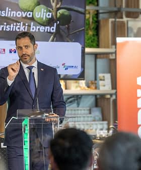 Bir etkinlikte kürsüde zeytinyağının bir turizm markası olarak tanıtımı hakkında konuşan takım elbiseli bir adam. Olive Oil Times