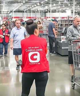 Женщина в красном жилете стоит в кассовой зоне магазина Costco на фоне тележек и покупателей. Olive Oil Times