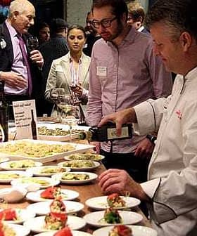Un chef verse de l'huile d'olive sur une assiette de nourriture lors d'un événement culinaire avec les participants qui l'observent. Olive Oil Times