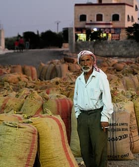 Un homme âgé portant une tenue traditionnelle se tient parmi des sacs en toile de jute remplis d'un produit non spécifié. Olive Oil Times